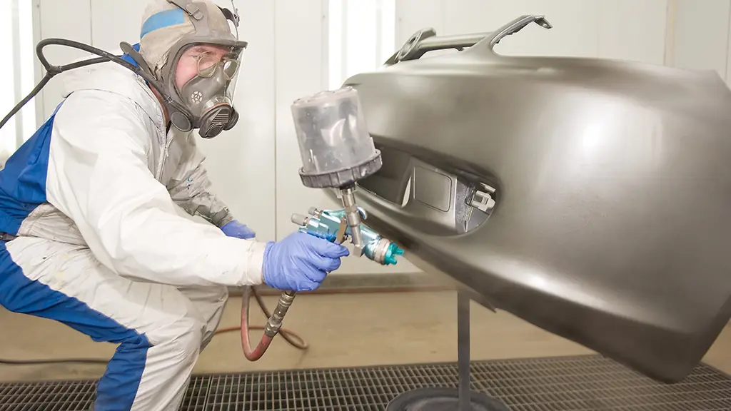 A technician in full respirator gear refinishing a bumper in the booth